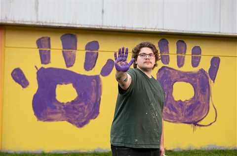 This is a photograph of artist Nicholas Currie in front of his mural titled Hands of Hurstbridge.