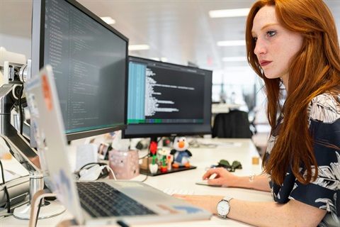 A person sitting at a desk and looking at two computer screens while holding a mouse