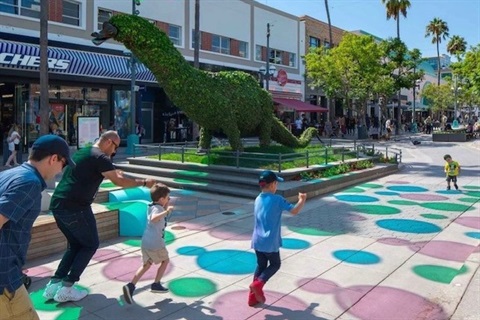 Photo of an outdoor shopping mall. A large green dinosaur sits in the middle of the street as children play nearby 