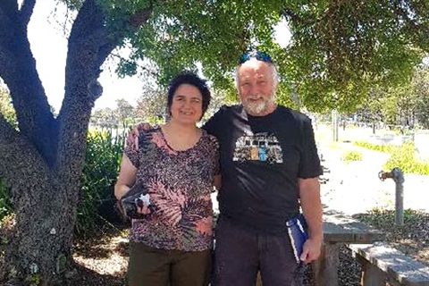 Two people stand under a shady tree, smiling.