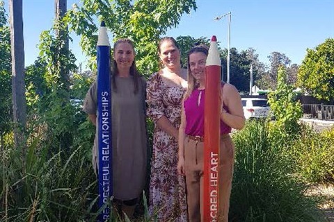 Women stand in a garden next to large pencils with the words 'Respectful Relationships' and 'Sacred Heart Primary School'