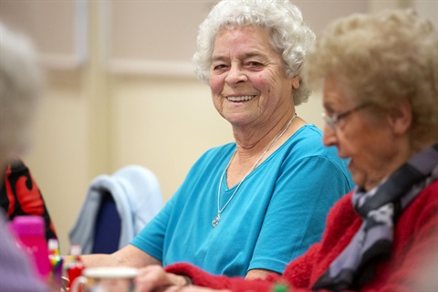 An older woman is sitting in a group, she's smiling at the camera