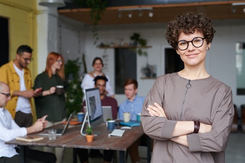 Photo of a woman standing with her arms crossed, smiling. Behind her a group of five people are in a group around a table, doing work.