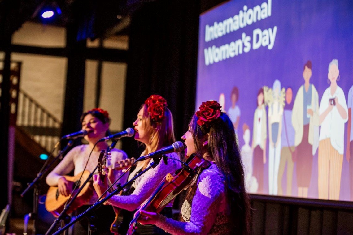 An all-female trio plays music in front of an International Women's Day sign at Eltham Community and Reception Centre in 2024