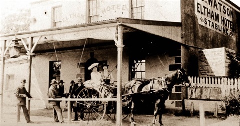 A historical photo showing two women in a horse-drawn cart in front of a building. The side of the building reads 