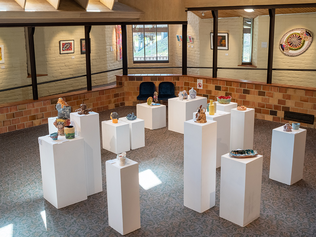 Image of colourful small sculptural works installed on different shaped plinths in the middle of the gallery, framed works hung on the mudbrick walls by Araluen Studio Artists