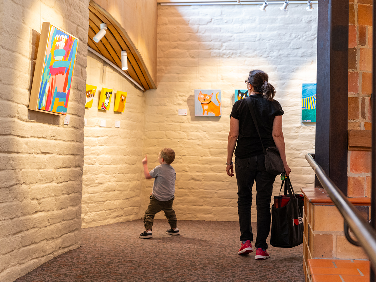 Colourful and playful paintings of domestic settings and pets installed on the gallery walls, with a family viewing the paintings.