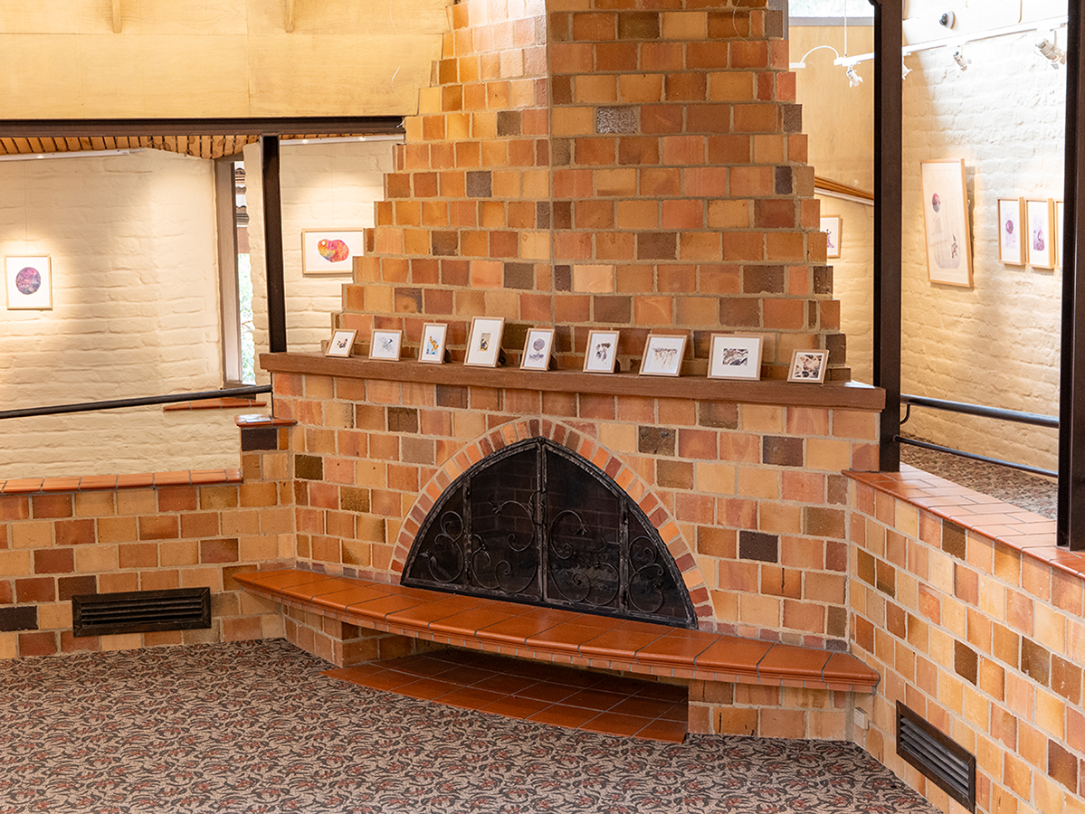 Photo of framed works installed on a ledge on a brick fireplace and on the gallery mudbrick walls