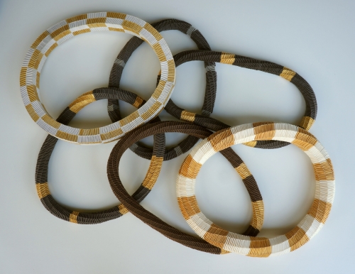 Visual description: Six woven necklaces overlapping each other on a white background. The necklaces have blocks of brown, gold, white and black colours.