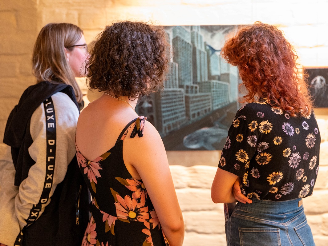 A photo of three people standing and looking at a painting of a dark cityscape