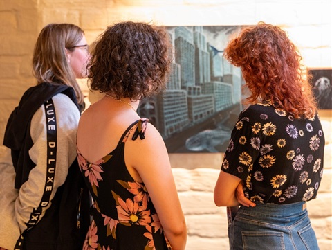 A photo of three people standing and looking at a painting of a dark cityscape