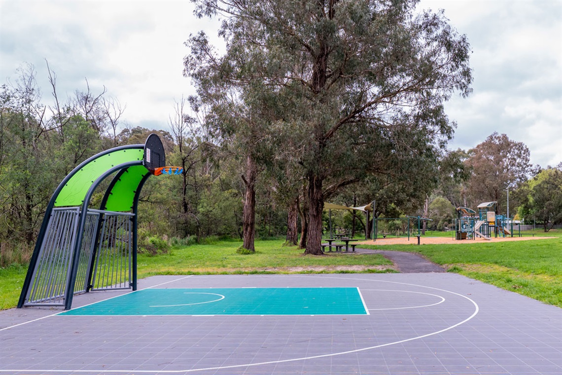 A basketball half court sits in front of a large tree and children's playground.