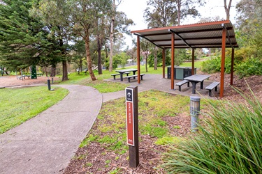 A shelter with tables and chairs sits next to a trail. There is a sign to Bunjil Reserve next to the trail.