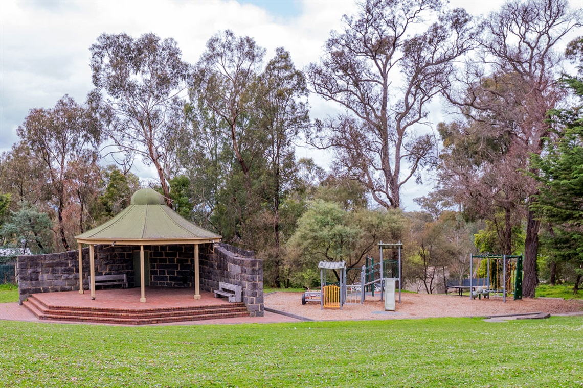 An old style rotunda sits on a paved area, it's next to a small playground.