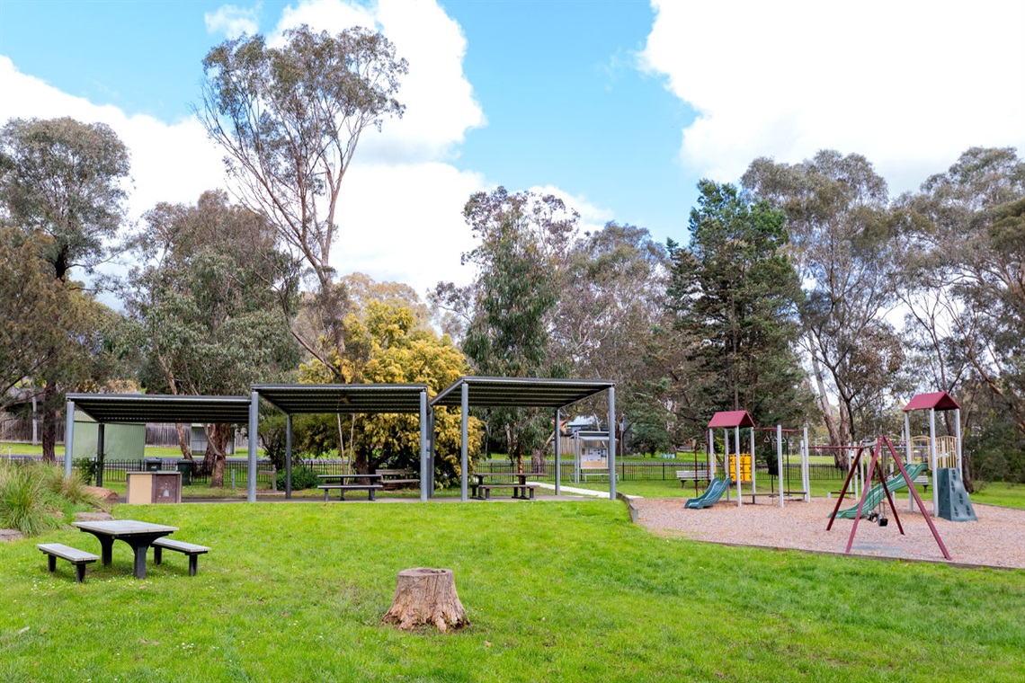 Shaded areas with picnic tables and a children's playground sit in front of trees and behind a large green grassed area. 