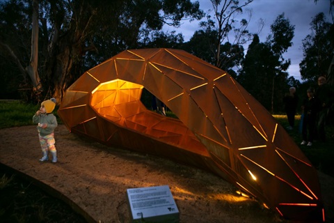 This is a photograph of the Eltham Gateway sculpture lit up in the early evening with a toddler standing in front of it.