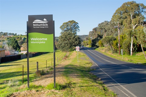 Photo of a bitumen road with grass and trees on either side. There is a sign that says 