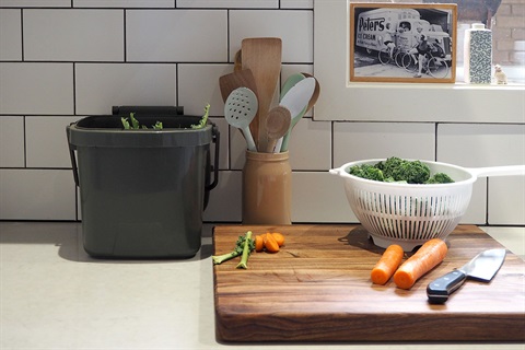 A kitchen bench has a chopping board, food preparation and a grey plastic bin