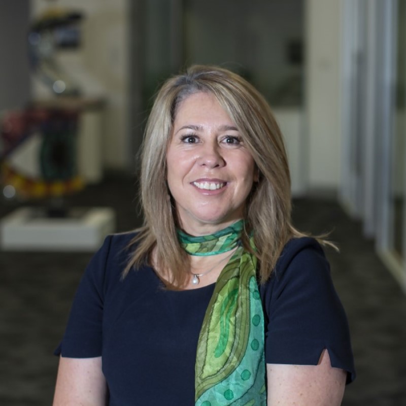 Photo of Corrienne Nichols. Corrienne is wearing a black shirt and green scarf and standing in the foyer of the Nillumbik Council building.