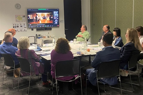 A group of adults are gathered around a meeting table looking at a monitor on the wall