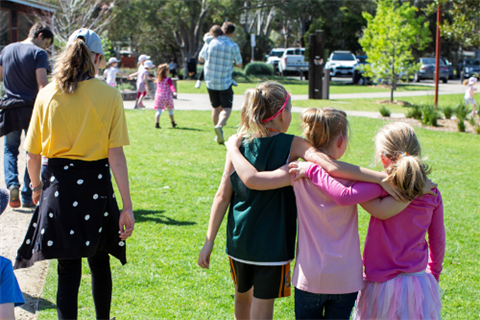 Three young girls walking arm in arm, other people of different ages all walking across a green lawn. 